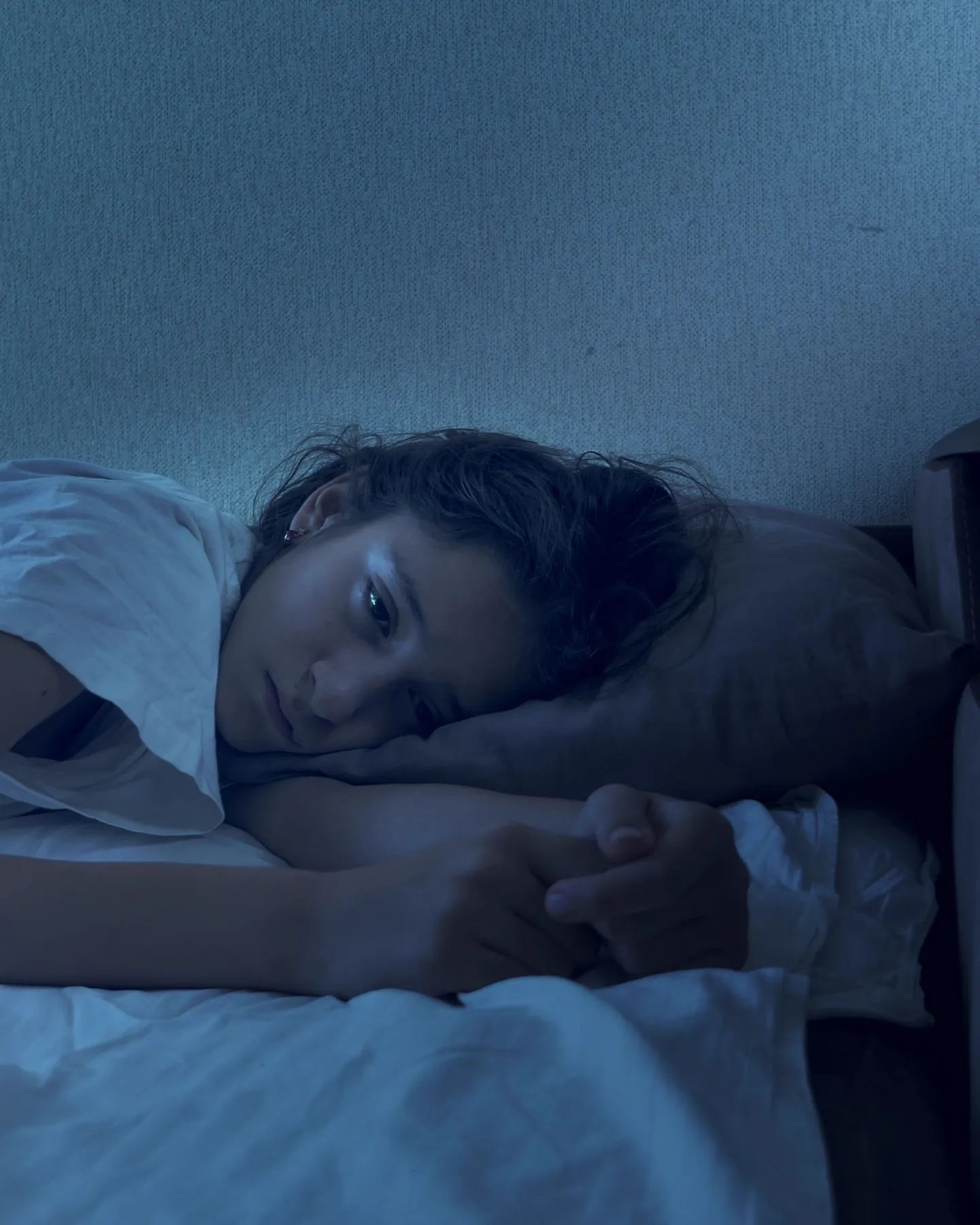 A teenage girl lies awake in bed at night, her eyes open in the dim blue light and her hands drawn up under her chin.